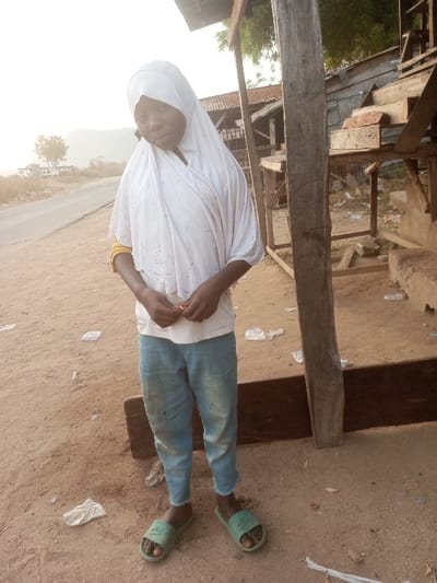 This full-length portrait captures a young person, likely a child or pre-teen, standing outdoors in what appears to be a rural or semi-urban setting in Ebiya, Nigeria. The subject has dark skin and is dressed in a modest white hijab that covers their head and shoulders, a white short-sleeved top, and light blue, worn jeans. They wear green slide sandals on their feet. Their hands are clasped in front, holding a small, light-colored object. The person is looking towards the camera with a neutral, slightly thoughtful expression.
The scene depicts a dusty, unpaved ground littered with crumpled plastic bags and debris. To the left, a paved road stretches into the background, with sparse trees, distant hills, and structures under a bright, somewhat hazy sky, suggesting early morning or late afternoon light. To the right of the subject, there are simple, rustic wooden structures, possibly part of a market stall or an outbuilding, with a prominent wooden pole standing vertically near the center-right of the frame. The overall environment conveys a sense of everyday life in a modest, outdoor location. There is no visible text in the image.