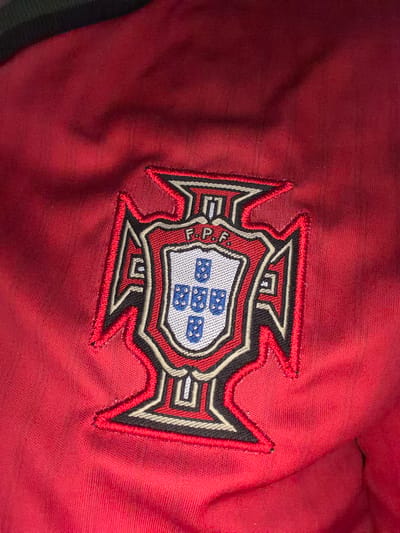 The image shows a close-up of an embroidered emblem on a red fabric. The emblem is shield-shaped with ornate borders and features a smaller shield in the center. The central shield contains five blue escutcheons, each with five white bezants. Above the central shield, the letters "F.P.F." are visible in white embroidery. The outer shield is outlined in red, black, and white embroidery, with stylized angular shapes extending outwards. The red fabric appears to be a garment, possibly a jacket or shirt, with visible stitching and texture. The lighting in the image creates some shadows and highlights on the fabric, emphasizing its texture and the dimensionality of the embroidery. The background is out of focus, showing more of the red fabric. No specific location in Nikopol, Bulgaria, can be determined from this image alone.