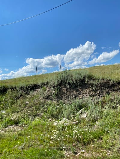 This is a vertical, medium-range outdoor shot of a grassy hillside with a blue sky and white clouds above, taken on a bright, sunny day. A thin, dark wire stretches diagonally across the upper portion of the frame, supported by two poles that are visible on the hillside. The hillside itself is covered in green and yellow grasses, with patches of bare earth and rocks. Small white wildflowers are scattered throughout the foreground. The scene appears to be a natural, undeveloped area. The location is identified as Yuldybayevo, Russia.