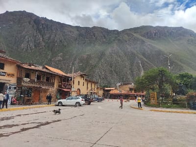 This image captures a bustling street scene in Ollantaytambo, Peru, set against a dramatic backdrop of towering, rugged mountains under an overcast sky. The foreground features a wide, paved town square or main street, showing dark, damp patches, suggesting recent rain or wet conditions.

On the left side of the street, a row of traditional, multi-story buildings stands out with their warm ochre and brown facades, rustic wooden balconies, and distinct terracotta roof tiles. These structures appear to house various establishments, with visible text on signs, though partially obscured, including "TOURS" and what seems to be "ARTESANIAS" (handicrafts), indicating local businesses. A white SUV (possibly a BMW) and a darker utility vehicle are parked along the curb in front of these buildings.

Several people are scattered throughout the scene, engaged in everyday activities. On the far left, a few individuals are standing near the storefronts. In the middle of the street, a small dog with dark fur ambles across the pavement. Further to the right, two people are walking: one wearing a distinctive yellow safety vest, and another in a striped shirt, both moving towards the center of the square. On the right side of the square, a verdant park area is visible, featuring lush trees, shrubs, and a dark-colored statue, possibly equestrian, adding a historical or cultural element. A tall, multi-armed streetlamp stands prominently within this park area. The mountainous backdrop, covered in sparse green vegetation, dominates the upper half of the image, emphasizing the dramatic natural setting of this historic Peruvian town. The time of day appears to be daytime, under a moderately cloudy sky, creating a soft, even light across the scene. The overall impression is one of a peaceful, functional town blending local life with a majestic ancient landscape.