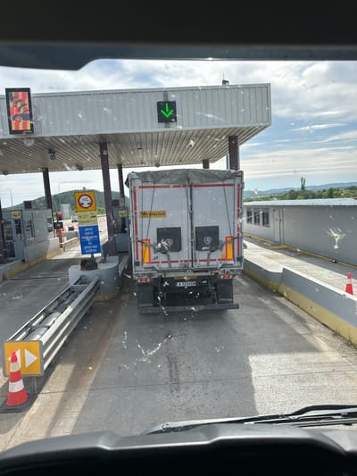 The image shows a truck passing through a toll booth. The truck is white with red and grey markings and the words "WIELTON" written on it. The license plate of the truck is A 5214 EM. The toll booth has signs indicating a height restriction of 10 meters and that all Greek tags are accepted. The location is likely in or near Αρδάνι, Greece, based on the Greek language on the signage and the "Egnatia Pass" sign. The toll booth is white with a green arrow indicating the lane is open. There are other vehicles waiting in line behind the truck. The view is from inside a vehicle.