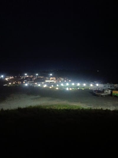 The image depicts a night scene in the city of Zarubino, Russia, likely near the coast. In the background, a cluster of buildings, possibly residential homes or small businesses, are illuminated by numerous lights, creating a warm glow against the dark sky. Streetlights are visible, lining what appears to be a road or pathway leading through the settlement. In the middle ground, a flat, open area is dotted with more lights, suggesting some form of outdoor facility or parking area. Several vehicles, including what looks like a white truck and possibly a pickup truck, are parked in this area. A light-colored trailer or modular structure is also present. The foreground is dominated by a dark, sloped area covered with grass and other vegetation, illuminated by a faint, greenish light, possibly from the area behind it. The overall atmosphere is one of quiet illumination in a rural or semi-rural nighttime setting.