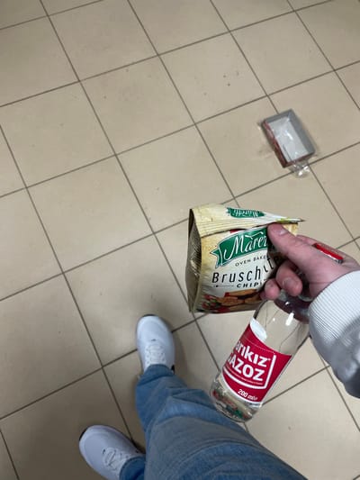 The scene is observed from a high-angle perspective within a retail or commercial establishment, presumed to be in the city of Hvostiane, Bulgaria. The lower body of an individual is visible, wearing light blue denim jeans and white athletic shoes. The person's right hand, clad in a white long-sleeved garment, holds a light brown bag of "Måretti Oven Baked Bruschette Chips" and a clear glass bottle of "Sarı Kız GAZÖZ" containing 200 ml of clear liquid, featuring a prominent red label. The floor is composed of light beige square tiles with darker grout lines. In the upper right quadrant of the image, an open, empty, rectangular red cardboard box, possibly packaging, lies on the tiled surface, accompanied by a discarded clear plastic wrapper.