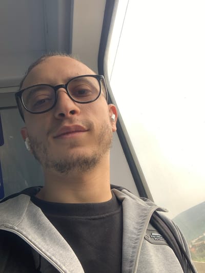 A male individual is depicted in a selfie, positioned indoors within what appears to be a vehicle in or around the city of Sidi Kacem, Morocco. He has light skin, a receding hairline, and a short, dark beard. He wears black-framed eyeglasses and white wireless earbuds in both ears. His attire includes a black crew-neck long-sleeve shirt underneath a grey zipped hoodie, which bears a "Mercedes-AMG Petronas Motorsport" logo patch on the upper right chest. The individual looks directly into the camera with a neutral expression. To his right, a vehicle window shows a bright, overcast sky, with a sliver of a distant green and brown hillside, possibly containing structures, visible in the lower right corner. The interior upper paneling of the vehicle is visible in the top left of the frame.