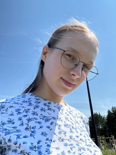 A low-angle, medium shot captures a young woman wearing round, wire-frame glasses and a light blue dress with a floral print, outdoors under a clear sky. Her hair is pulled back, with some strands blowing in the wind. The background shows a hint of a forest and a playground structure. The woman's face is tilted slightly downwards, and she is smiling faintly. The image is likely taken in Металлплощадка, Russia, given the context.