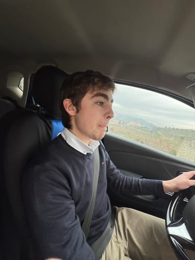 The image displays the interior of a vehicle, focused on a male driver and the exterior landscape through his window. The driver, a young adult male with light skin, short brown hair, and light facial stubble, occupies the left side of the frame. He is dressed in a white collared shirt, a dark blue crew-neck sweater with a small red, white, and blue emblem on the left chest, and khaki trousers. A dark gray seatbelt is fastened across his torso and lap. His left hand rests on the steering wheel, which is black with reflective accents. His mouth is slightly open, and his gaze is directed forward-right, towards the exterior. The vehicle's interior features a dark grey headliner, seat upholstery, and door panel. Through the driver's side window, an expansive hilly landscape is visible under an overcast sky. The terrain is characterized by sparse green and brown vegetation, with distant buildings and cultivated plots indicative of an urban-rural interface. This scene is situated in the city of Lamego, Portugal. The vehicle appears to be in motion.