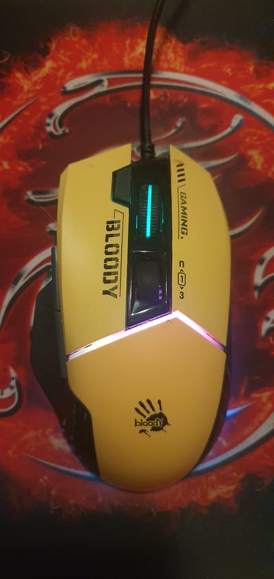 The image shows a close-up, overhead view of a yellow and black computer mouse with RGB lighting. The mouse is resting on a red and black patterned mousepad. The mouse has the brand name "BLOODY" printed vertically on its left side. On the right side, there are markings that appear to be related to gaming functions, including "GAMING" and a numerical indicator. The scroll wheel is illuminated with a bright blue light, and there is a purple glow emanating from the base of the mouse. The background mousepad features an abstract fiery or lava-like design with silver accents. There are no people or any other identifiable objects or environmental features visible. The location in Alūksne, Latvia, is not discernible from the image.