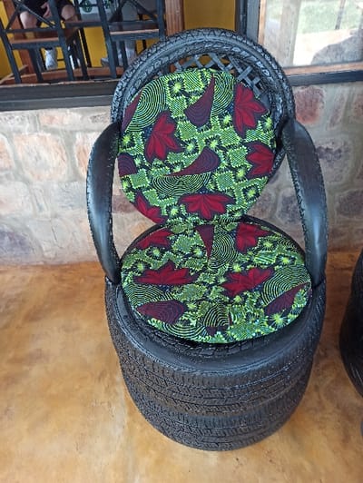 Two chairs made from stacked car tires, painted black, are side-by-side on a tan floor. The seats are fitted with round cushions covered in a colorful fabric featuring green, yellow, and red floral patterns. Each chair has a curved backrest also fashioned from a tire. They sit near a wall composed of light-colored stones and a window.