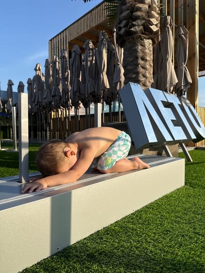 A young child, viewed from behind and in profile, is kneeling on a rectangular, light-colored raised platform covered with artificial grass. The child is wearing patterned swim shorts with a green and white checkerboard design and a yellow circular emblem. The child's head is bowed and resting on their arms, which are extended forward on the platform. In the background, there is a large, reflective, metallic sign with the letters "NETO" in capital letters, tilted backwards. Behind the sign and the child, rows of folded gray beach umbrellas are visible, along with a palm tree and a wooden structure with vertical slatted walls and a corrugated roof. The setting appears to be an outdoor recreational area, possibly a beach club or resort, under a clear blue sky. The location is identified as Dumino, Russia.