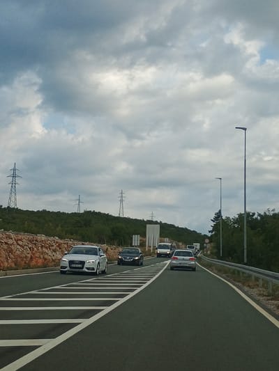 This is a vertical, eye-level, medium shot of a highway in Praputnjak, Croatia, under a cloudy sky. In the foreground, the asphalt road is marked with white lines. Several cars are driving on the highway, including a white Audi A3 in the left lane, a black sedan in the right lane behind the Audi, and a silver Volkswagen Golf further down the right lane. In the distance, more vehicles are visible, including vans and cars. The highway is bordered by a metal guardrail on the right and a rocky embankment and green vegetation on the left. In the background, the landscape is covered in dense green trees and bushes, with several high-voltage electrical towers visible on the hills. A streetlamp is present on the right side of the road. The sky is filled with dramatic, gray clouds, indicating overcast weather.