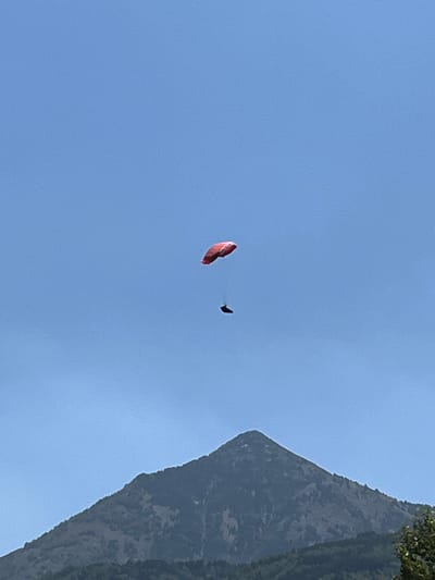 In Sahún, Spain, a paraglider, identified by a red canopy and a dark figure beneath it, is captured mid-flight against a clear blue sky. Below the paraglider, the rugged, tree-covered slopes of a mountain dominate the lower portion of the frame, with the peak of the mountain reaching towards the paraglider. The scene suggests an outdoor recreational activity taking place in a mountainous region.