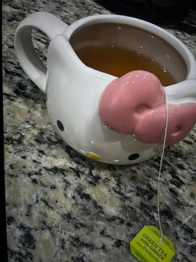 A white, ceramic Hello Kitty-themed mug, featuring black eyes, a small yellow nose, and a prominent pink bow on its front, is positioned on a light-colored, mottled granite or quartz countertop in the city of Arthès, France. The mug contains an amber-colored liquid, identified as tea, in which a Bigelow brand tea bag is steeping. A white string extends from the mug to a bright green tea tag labeled "BIGELOW GREEN TEA with GINGER plus Probiotics". The scene is static, devoid of human presence or overt action, focusing solely on the steeping tea.