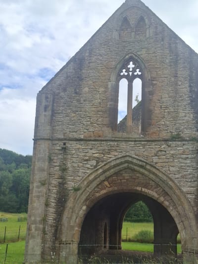 This is a vertical, eye-level shot of the ruined stone structure of a building, possibly a church or abbey, in Catterick Garrison, United Kingdom. The weathered stone blocks form a wall with a large arched opening on the right and a gothic-style window on the left. The window is intricately carved with a quatrefoil design in the upper section. Through the arched opening, a green field with a fence is visible, suggesting an outdoor setting with a rural or park-like atmosphere. A dense line of trees forms the horizon behind the field. The sky above is cloudy with patches of blue, indicating daylight but not necessarily bright sunshine. There is no visible text in the image. The overall impression is one of historical decay and the integration of architectural ruins with the natural landscape.