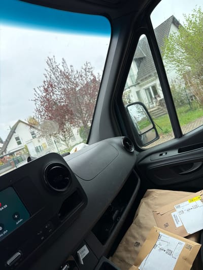 The image shows a view from inside a vehicle, likely a van, with the dashboard and front windows visible. The windshield shows a partial view of buildings and trees, suggesting a residential or suburban area. There are packages on the dashboard in the front right corner of the van. The packages are labeled with "Altkönigstraße 25" which indicates the location in Maintal, Germany. The image captures a moment inside the vehicle, potentially while parked or stationary. The dashboard features an infotainment screen displaying information and possibly navigation details. The overall setting seems mundane, indicative of a routine drive or stop in a residential area.