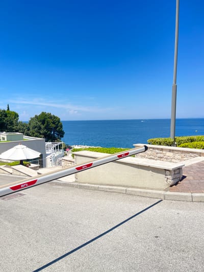 The image captures a sunlit outdoor scene with a clear blue sky and the Adriatic Sea in the background. A white barrier arm with red "Bft" logos is positioned diagonally across the foreground, obscuring part of a paved road. Beyond the barrier, a modern building complex with white and grey facades, lush green trees, and a white umbrella are visible. The coastline features stone accents and manicured hedges. In the distance, several small boats are scattered across the calm blue water. The location is identified as Crveni Vrh, Croatia.