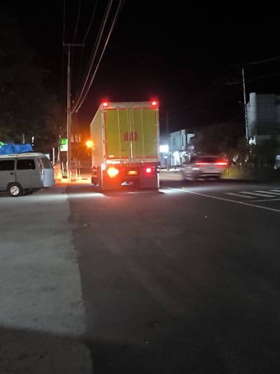 The image depicts a nighttime scene on a street in Paya Tringgadeng, Indonesia. A large, yellow and white truck with its brake lights illuminated is stationary in the right lane of the road. To the left of the truck, a gray van with a blue tarp on its roof is parked on the side of the road. A white car is visible in the distance, moving away from the viewer in the right lane. The street is paved, with a distinct line dividing the light-colored pavement on the left from the darker pavement on the right. Overhead, power lines are strung between utility poles. Buildings and trees line the background. The scene appears to be near a gas station, indicated by the presence of illuminated green signage in the background.