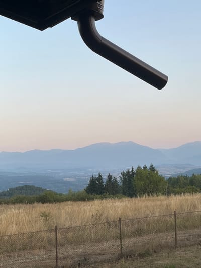 This image depicts a panoramic view of a rural landscape from an elevated position, identified as Gorni Okol, Bulgaria, under soft twilight conditions. In the upper left foreground, a dark, angled downspout and gutter system partially extends into the frame. The lower foreground features a segment of a chain-link fence with two upright metal posts, positioned across dry, sparse earth. Behind the fence, a wide expanse of tall, dry, golden-brown grass dominates the mid-ground. Beyond the grassland, a dense cluster of green evergreen and deciduous trees marks the edge of a forested area. The background reveals a wide valley or plain containing a discernible distant settlement with clustered buildings. Further in the background, multiple layers of hazy, blue-tinted mountains stretch across the horizon. The sky exhibits a clear gradient from light blue at the zenith to a faint pinkish-orange hue near the horizon. No people, subjects, or actions are present.