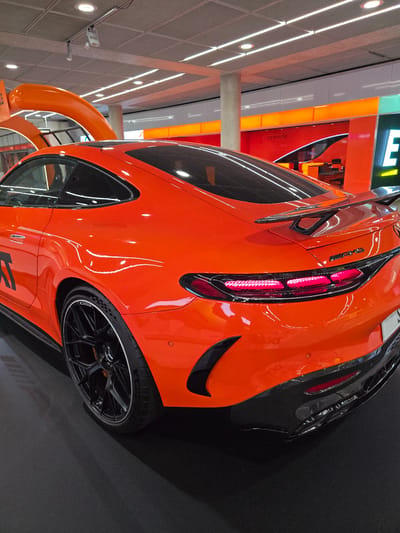 A bright orange Mercedes-AMG GT sports car is parked on a black floor. The car is viewed from the rear, with its sleek lines and aerodynamic features prominently displayed. The car has black rims and a large black spoiler. The background is blurred, but it appears to be an indoor exhibition space with orange and white lighting. Part of a large orange structure, resembling a curved ramp, is visible in the upper left quadrant of the image. In the upper right, a large white "E" logo is visible on a green background. The image appears to be taken at an event related to automotive or technology, possibly at Messe Stuttgart airport, located in Echterdingen, Germany, which is a convention center adjacent to the Stuttgart Airport.