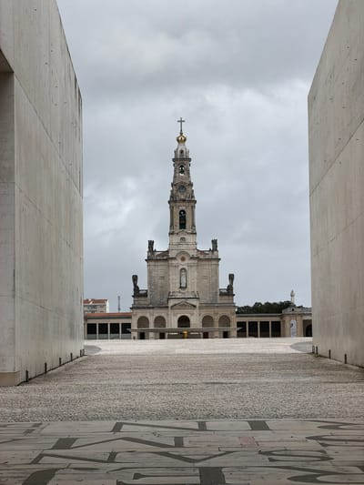 The image captures a long-distance view of the Sanctuary of Fátima in Fátima, Portugal, specifically the Basilica of Our Lady of the Rosary, under an overcast sky. The scene is framed by two prominent, smooth, light-colored concrete walls on the immediate left and right, creating a narrow, symmetrical aperture.
In the foreground, the ground is paved with large, irregularly shaped light-grey flagstones featuring dark, abstract, geometric patterns. This transitions into a vast, open plaza paved with uniform light-grey cobblestones, extending towards the basilica.
The Basilica of Our Lady of the Rosary stands centrally in the middle ground, a grand stone edifice with a dominant central bell tower. The tower is multi-tiered, features a clock face, and is topped with a gilded crown-like structure surmounted by a Latin cross. The main façade of the basilica below the tower includes a central statue of the Virgin Mary in a niche, flanked by architectural details and additional statues. The basilica's base incorporates a series of open arches forming a loggia. To the left and right of the basilica, extending horizontally, are colonnaded walkways or buildings, part of the broader sanctuary complex. A smaller, modern building with an orange-tiled roof is visible behind the left colonnade. To the far right, beyond the right colonnade, a white statue on a pedestal is discernible.
A single individual, dressed in a bright yellow or lime green garment, is observed standing on the cobblestone plaza directly in front of the basilica's arched entrance, appearing to face away from the viewer. No other individuals are distinctly visible. The sky is entirely covered with thick, light grey clouds, suggesting an overcast day with diffused lighting.