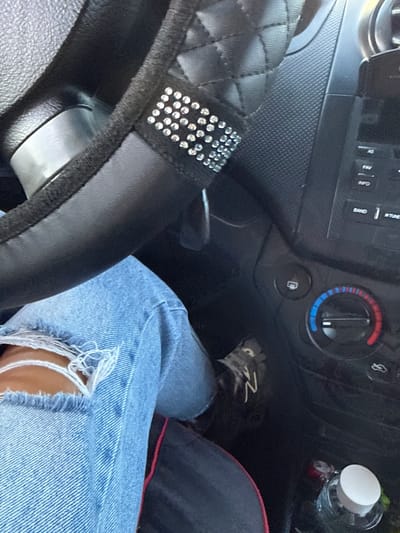 The image shows a close-up view of the interior of a car, specifically the steering wheel area and a person's lap. A person's legs, clad in ripped light-wash denim jeans, are visible. The steering wheel is black with a diamond-stitched pattern and embellished with rhinestones. Part of the car's dashboard is also visible, including climate control dials and buttons for the car's audio system. A bottle of water with a white cap sits on the floor in the lower right corner. The context of Sadaclia, Moldova is not discernible from the image itself.