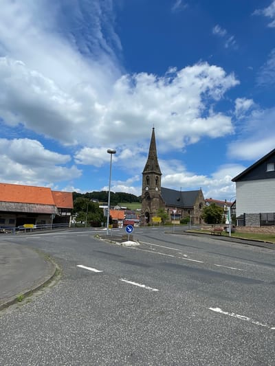 The image shows a view of a street intersection in Seigertshausen, Germany. In the background, a prominent church with a tall, pointed steeple is visible. The church appears to be constructed of stone and has a dark roof. To the left of the church, there is a building with a red tiled roof and half-timbered details. To the right of the church, a modern building with white brick and a dark roof is present. The intersection itself is paved with asphalt and marked with white lane lines. A street lamp stands on the left side of the road, and a blue directional sign with a white arrow pointing downwards is positioned on the right. There are also a few trees and green spaces visible in the scene, along with a bench on the right side of the road. The sky is blue with scattered white clouds.