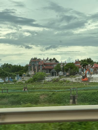 The image shows a cemetery in Hải Lăng Commune, Vietnam. The sky is overcast with heavy, gray clouds. In the foreground, there is a grassy area with some fencing and a concrete barrier. Behind this, the cemetery is visible with its elaborate tombs, some of which have red tiled roofs and ornate decorations. Several trees and lush green vegetation surround the tombs. An orange excavator is parked to the right of the cemetery, suggesting ongoing construction or maintenance. The overall scene is somber, characterized by the cloudy sky and the presence of a cemetery.
