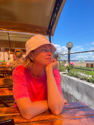 A woman wearing a pink t-shirt and a white bucket hat sits at a wooden table outdoors, resting her chin on her hand and looking to the right. She has blonde hair and is wearing small hoop earrings. Behind her, a man and a woman are seated at another table. In the background, there is a beach with a playground, a body of water, and a clear blue sky with clouds. The overall setting appears to be a casual outdoor dining area near a beach, possibly in the city of Belozersk, Russia, given the context.