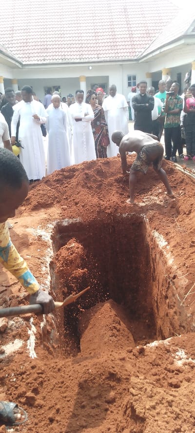 This media file depicts an outdoor scene, likely a burial or grave preparation, taking place in Umu Enge, Nigeria, under natural daylight conditions.
The foreground is dominated by a freshly dug, rectangular grave in reddish-brown earth, with a large mound of the same soil piled next to it. In the bottom left, a person is seen from the chest down, wearing a patterned long-sleeved shirt, actively using a shovel to move soil into the grave. To the right, another individual, shirtless and wearing patterned shorts, is bent over, appearing to be working within or on the edge of the grave, possibly shaping or clearing it with their hands. White powdery residue, potentially lime, is visible on the edges of the grave and mixed with the soil.
In the background, a group of people stands observing the activity. Prominently featured are several men dressed in long white clerical robes, suggesting they are priests or religious leaders. One man in a black clerical shirt with a white collar is also visible. Other attendees wear various forms of dress, including one woman in a patterned traditional outfit and head covering. Their collective presence indicates a gathering for a solemn occasion.
Behind the observers, a building with a light-colored tiled roof and white walls is visible, possibly a church or a public hall, completing the setting. The overall image captures a somber event centered around the interment process. No visible text is clearly discernible within the image.