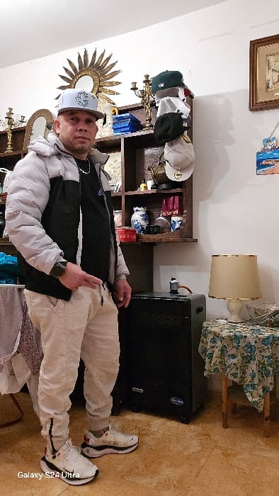 This image depicts an adult male, estimated to be in his mid-thirties to forties, standing centrally within an indoor residential setting. He is positioned facing directly forward. The individual wears a light grey baseball cap with a white logo, a silver chain necklace, a black t-shirt, a black and light grey puffer jacket, light beige cargo-style pants with black stripe detailing on the lower legs, and white sneakers with black accents and visible "GORE-TEX" branding. A dark smartwatch is present on his left wrist. His right hand holds a drawstring from his pants, while his left hand is positioned near his left hip.
The room features white walls and a light-colored, mottled tiled floor. In the background, a dark wooden multi-shelf unit displays various decorative items. These include a large golden sunburst-style mirror mounted on the wall above the unit, a golden candelabra, multiple baseball caps (green, black, beige, and a white/grey hat) hanging or resting on the shelves, blue-bound objects, blue and white patterned porcelain vases, red cylindrical containers, and various glass bottles. To the immediate right of the individual, a large black speaker box on wheels is situated on the floor. Further right, a small wooden stool or table with a blue and white floral patterned tablecloth holds a cream-colored table lamp with a gold-trimmed shade and a small silver metal object, possibly a coffee maker. Two framed artworks are visible on the wall above this table: one depicting buildings and another a coastal scene. A fabric-draped furniture item is partially visible on the far left. The bottom left corner of the original image contains a "Galaxy S24 Ultra" watermark. The specific city of Real Sitio de San Ildefonso, Spain, cannot be confirmed from the visual information within the image.