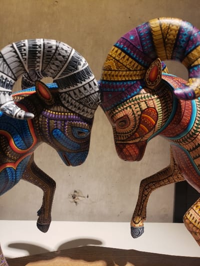 Two intricately decorated wooden ram figurines are stacked vertically. The top ram displays vibrant colors including yellow, orange, purple, and turquoise with detailed patterns. The lower ram features contrasting black and white patterns on its horn, while its body is adorned with blue and purple designs. The background includes a pale wall with visible texture and a partial view of a wooden structure on the right side.
