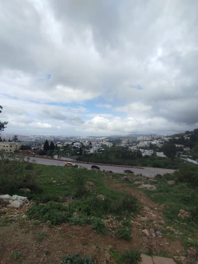 The image captures a wide-angle view overlooking the city of Tétouan, Morocco, from an elevated hillside perspective. The scene is set under an overcast sky, dominated by large, textured grey and white clouds, with patches of clear blue sky visible in the upper mid-frame, suggesting breaks in the cloud cover.

In the immediate foreground, the terrain is rough and unkempt, with dry, reddish-brown dirt, scattered rocks, and patches of green, bushy vegetation. Some white plastic bags and other debris are visible on the ground, indicating litter.

The mid-ground features a paved road, likely a two-lane thoroughfare, curving gently from left to right across the frame. Two cars are visible on the road: a bright yellow car, possibly a taxi, and a dark-colored car following it. The road is bordered by more green, grassy hillsides dotted with trees and light-colored buildings, characteristic of residential areas. These buildings, mostly white or off-white with flat roofs, are nestled into the undulating landscape.

In the distance, the urban sprawl of Tétouan extends across a vast plain, with numerous buildings stretching towards a hazy horizon where faint outlines of mountains or distant hills are visible. The overall impression is that of a bustling city expanding into its surrounding natural environment.

The time of day appears to be daytime, with soft, diffused light, typical of an overcast condition, though the visible blue sky suggests sunlight might be breaking through intermittently. No people or visible text are discernible in the image.
