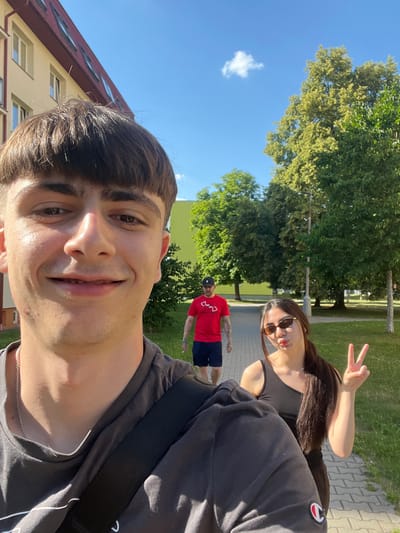 This is a selfie taken outdoors on a sunny day, likely in Dobřany, Czechia. The camera is held at a low angle, capturing the faces of two young people in the foreground and background. The main subject, a young man, is in the immediate foreground, with his face close to the camera and a wide smile. He has dark, short hair and is wearing a dark t-shirt and a silver chain.

Behind him, a young woman is also looking at the camera, wearing sunglasses and a black tank top. She is making a peace sign with her fingers. In the middle ground, another person wearing a red t-shirt and dark shorts is visible, facing away from the camera. The background features lush green trees, a paved walkway, and a yellow building with a red roof. The sky is a clear, bright blue with a small white cloud. The overall mood appears to be cheerful and relaxed, suggesting a casual outing or gathering. The sunlight indicates it's daytime.