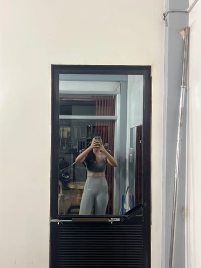 A person is taking a mirror selfie in what appears to be a gym. The person is wearing a grey sports bra and grey leggings. In the reflection, a gym environment is visible with equipment and other people. The overall scene suggests an indoor fitness setting. The provided location of Cuyo, Philippines, is an island municipality in the province of Palawan.