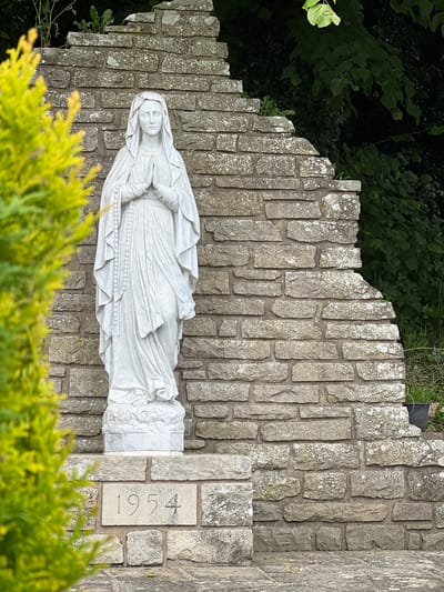 This media file displays a vertical orientation. The main subject is a white marble statue of the Virgin Mary, adorned with a rosary. The statue is positioned against a stone wall constructed from irregular grey stones. To the left of the statue, a light-colored stone slab with the inscription "1954" is visible, indicating a potential date of construction or dedication. The scene is partially obscured by lush green foliage at the top and bottom, suggesting an outdoor setting, possibly a garden or memorial. The lighting appears natural, hinting at daytime, and the overall atmosphere is serene and contemplative. The location context is Mountcharles, Ireland.