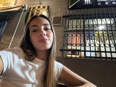 A young adult female is depicted in a close-up, selfie-style photograph, positioned in the foreground and looking directly at the camera with a slight smile. She has long, light brown hair, a small piercing on her left eyebrow, and wears a plain white t-shirt. The background reveals the exterior of a building at night in Plasencia, Spain. A prominent sign on the upper left reads "EL ESQUINAZO PLASENCIA," partially obscured by a dark awning. To the right, a window is fitted with decorative black wrought iron security bars; reflections of interior lights and indistinct framed items are visible through the glass. Above the window, to the left, an outdoor air conditioning vent is mounted. A partial sign on the upper right shows "M **** CERA." The building's wall has a textured, light-colored finish.