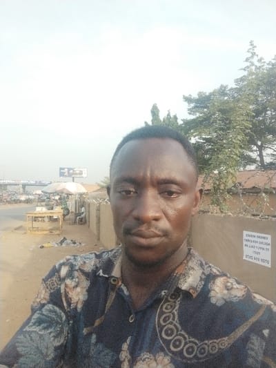 The image shows a man taking a selfie in front of a low brick wall with barbed wire. The man is wearing a blue and brown shirt with a floral pattern. The background shows a dusty street, a small roadside stall, and a billboard in the distance. The scene is located in Maraba, Nigeria, with coordinates of latitude 9.024 and longitude 7.573. On the wall behind the man is a sticker which says "0708 849 2475, TEXT TO APPLY CALL ON WSC  PER-EVENT, 125,000.000, WORKERS NEEDED".