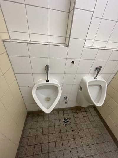 This is a forensic, matter-of-fact description of an image taken in a public restroom. The image shows two white ceramic urinals mounted on a white tiled wall. The urinals are of standard design, with a visible drain at the bottom of each bowl. The left urinal appears to have some debris in its bowl. A metal flush valve is visible above the left urinal, and another metal flush valve, partially obscured by its activation lever, is visible above the right urinal. To the left of the left urinal, a vertical pipe extends from the wall. To the right of the right urinal, a small, dark object protrudes from the tiled wall, possibly a screw or nail. Below the urinals, the floor is covered with speckled grey tiles. A floor drain with a metal grate is located in the foreground, between the two urinals. The walls are tiled with white rectangular tiles, and the side wall visible on the left is tiled with a lighter, off-white color. The image does not contain any people or evidence of recent activity, other than the presence of debris in one of the urinals. The lighting appears to be artificial, providing a somewhat sterile environment. The image is set in a public restroom, likely a men's restroom, and is located in Bonn, Germany.