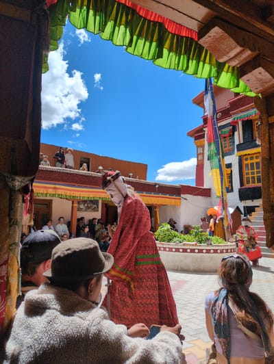 This photograph captures an outdoor courtyard in Pibiting, India, on a sunny day with a clear blue sky and scattered white clouds. The scene is framed by wooden beams and colorful, ruffled fabric draped across an overhead structure. In the foreground, several individuals are visible, including two people facing away from the camera, one wearing a light-colored, textured jacket and a hat, and another with a patterned scarf in their hair.
The central focus of the image is a person in elaborate ceremonial attire, including a red robe with green stripes and a white mask with red accents. This individual appears to be the subject of a cultural or religious event, with other people gathered to observe. Behind this figure, a multi-story building with white and red walls, adorned with colorful prayer flags and ornate yellow-trimmed windows, stands prominently. Further back, more spectators can be seen on balconies and in the courtyard. The ground is paved with interlocking stones. The atmosphere suggests a vibrant, communal gathering, likely a festival or ritual, taking place within a temple or monastery complex.