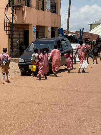 In Tangu, Nigeria, a group of children walks past a parked dark-colored SUV on a dusty road. The children are dressed in school uniforms, with some wearing pink dresses and others shorts and shirts. One child on the left, facing away from the camera, has a blue backpack with a stuffed animal attached. A few of the children are carrying yellow baskets, and one child is holding a white plastic bag. In the background, a light peach-colored building with a spiral staircase is visible, along with other structures and a few individuals in doorways. The sky is partly cloudy.