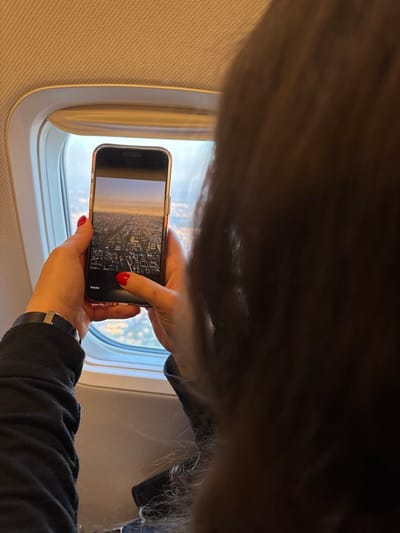 A passenger on an airplane holds a smartphone in front of a window, taking a photograph of the landscape outside. The phone's screen displays the image of a densely built urban area, likely a city, below. The passenger is wearing a black long-sleeved shirt and a wristwatch with a black band. Their nails are painted red. Part of the passenger's hair is visible in the top of the frame. The airplane window is white. The airplane interior has a beige wall lining. The location cannot be pinpointed to Vecsés, Hungary, based on the given image.