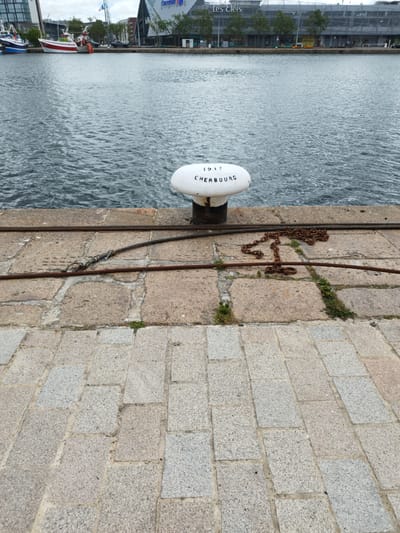 This is a waterfront scene in Cherbourg-en-Cotentin, France. In the foreground, a paved walkway made of granite slabs leads away from the viewer. A stone quay runs along the water's edge, with thick metal cables and a rusty chain lying on its surface. A white bollard, marked with "1917 CHERBOURG," stands near the water. The water is choppy and grey, reflecting the overcast sky. In the background, several boats are docked along the quay, including a white and red fishing vessel. Beyond the boats, a modern building with glass windows and a sign reading "Les Clés" is visible, indicating it is likely a commercial area. The overall impression is of a functional port area on a cloudy day.