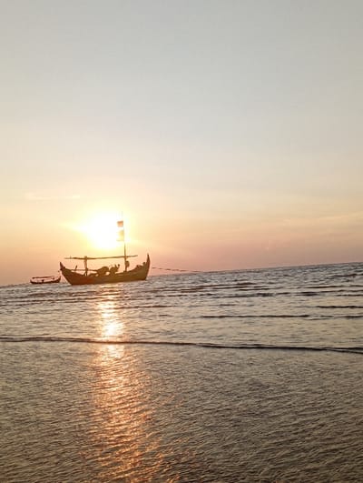 The image displays a coastal marine environment at sunrise or sunset in the city of Tritunggal, Indonesia. A brilliant, overexposed spherical sun is centered on the horizon line, emitting intense light. The sky above transitions from bright white-yellow at the horizon to pale orange and light blue-gray towards the top. The lower two-thirds of the image consist of calm water with gentle ripples, reflecting the sun's light in a prominent, shimmering golden path extending from the horizon to the foreground. In the mid-ground, a traditional wooden fishing boat is silhouetted against the sun. This boat features a high, curved bow and stern, and a prominent mast-like structure with horizontal beams. To the immediate left of the main boat, a smaller, narrower boat, also in silhouette, is visible. No distinct human figures or specific actions are discernible on or around either vessel.
