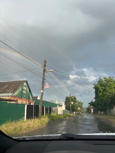 A double rainbow arcs across a cloudy sky over a wet, asphalt road in the city of Pokotylivka, Ukraine. The road is lined with green fences, houses, and trees. A utility pole with wires and a traffic sign depicting children is visible on the right side of the road. The scene appears to be captured from inside a vehicle, with the edge of a car's windshield visible in the foreground. The sky is dark and overcast, suggesting recent rain.