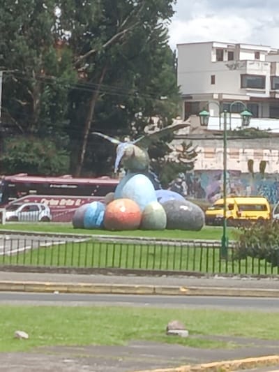The image displays a daytime scene at the Redondel del Águila (Eagle Roundabout), located at the intersection of Av. General Enríquez and Av. Luis Cordero in Sangolquí, Ecuador.
In the center of a grassy median, a prominent sculpture features an abstract bird-like figure with outstretched wings, predominantly in a light green hue, perched atop a cluster of large, smooth, rounded objects. These objects exhibit various colors, including light blue, red-orange, light green, dark blue, and dark gray/black. A low black metal fence encircles the sculpture and its immediate grassy base.
Surrounding the median are paved roadways. To the left of the sculpture, a large, dark red articulated bus is visible, with partial text "Orion B..." on its side. Behind the bus, a white compact car with a patterned black stripe on its body is present. Lush green trees form a dense background behind these vehicles.
To the right of the sculpture, a tall, dark green street light pole with two lantern-style fixtures stands in the midground. Beyond the streetlight, a multi-story, light-colored building with multiple windows and balconies is visible. A lower wall adorned with colorful graffiti stretches below this building. A yellow van is parked or moving in the background to the right. The sky overhead is partly cloudy. No discernible people or specific interactions are visible within the frame.