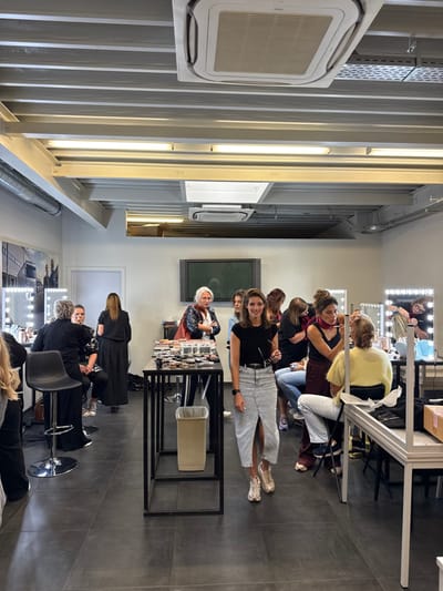 An indoor scene in a commercial or educational establishment located in Herentals, Belgium, depicts approximately ten to twelve individuals, primarily women, engaged in a makeup application or training session. The room features dark grey tiled flooring, white walls, and an industrial-style ceiling with exposed white beams, perforated panels, and rectangular fluorescent strip lighting, along with two white ceiling-mounted air conditioning units.

In the foreground, a light-skinned woman with dark hair, wearing a black short-sleeved top, a light-wash denim-style midi skirt with a front slit, and light-colored sneakers, walks towards the viewer while smiling. She holds sunglasses in her right hand.

To the left, a woman with short grey hair, dressed in black, sits on a black adjustable bar stool, applying makeup to another woman seated opposite her. Behind them, another woman in black stands partially obscured. A large monochrome poster depicting trucks and people hangs on the far-left wall.

In the midground, a black metal table is laden with various makeup products, including palettes and brushes, alongside a beige waste bin. Behind this table, an older woman with light hair, wearing a black leather-look jacket over a red garment, stands looking towards the left. A large, dark flat-screen television is mounted on the wall further back.

To the right, multiple makeup stations are set up, each featuring a white vanity table with a large rectangular mirror framed by circular vanity lights. A woman in a dark top and a red scarf is actively applying makeup to a person seated with their back to the camera, wearing a yellow fuzzy top. Further right, another woman with dark hair and glasses is seen in reflection within a vanity mirror, applying makeup to what appears to be a mannequin head or a person with light hair. Other individuals are present in the background, some seated, some standing, observing or participating in the activities. The overall environment suggests a professional or semi-professional setting for beauty or makeup instruction.