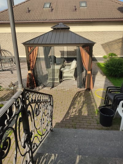 This is an outdoor scene in Vёшки, Russia, likely a residential backyard or patio area. The image features a brown-tiled roof of a building, a brick wall, and a paved patio area with interlocking brick pavers. A dark brown gazebo with retractable mesh sides and brown fabric curtains is centrally positioned on the patio. Inside the gazebo, there are white wicker chairs and a glass-topped table.
In the background, a stack of black plastic chairs and a black bucket are visible, along with a yellow garden hose. A lush green bush is situated to the left of the chairs and bucket. In the foreground, there is an ornate black metal railing with decorative scrollwork, suggesting a balcony or terrace edge. A lamppost with a decorative glass fixture is also present.
The scene appears to be bathed in sunlight, casting distinct shadows on the patio, indicating a sunny day. The shadows suggest the time of day could be late morning or afternoon. There is no visible text in the image. The overall impression is one of a well-maintained outdoor living space.