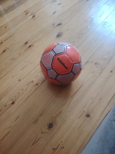A bright orange and gray soccer ball rests on a light brown wooden floor. The ball is slightly off-center, positioned in the lower right quadrant of the image, and is casting a subtle shadow to its left. The wooden floor is made of planks with visible wood grain and knots. The lighting suggests an indoor environment, possibly a home, with a bright light source reflecting on the floor from the upper right. There are no people or any other objects visible in the frame. The scene does not provide any information or context related to the city of Hovsan, Azerbaijan.