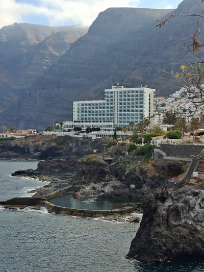 In Puerto de Santiago, Spain, a multi-story white hotel with blue-tinted windows is situated on a cliff overlooking the ocean. In the foreground, a natural swimming pool formed by rocks is visible, with waves crashing against the rocky coastline. A few people are present in the pool and on the surrounding rocks. The background features a steep, mountainous terrain with a hazy sky. The hotel and surrounding buildings are nestled on the hillside.