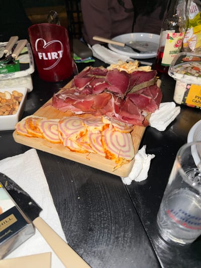 The image depicts a close-up view of a table laden with food and drinks, set in what appears to be a casual indoor gathering. In the foreground, a wooden cutting board holds a selection of cured meats, including thinly sliced prosciutto and pieces of darker, presumably aged, meat. Beside the meats, a rolled cheese product, sliced into rounds with a pink and white swirled pattern, is arranged. To the left, a white square dish contains a mix of nuts.

A dark red cup with a white heart and the word "FLIRT" is prominent in the upper left quadrant, slightly out of focus. Behind the food, a glass bottle with a red liquid, possibly a beverage, stands on the table. There are also clear plastic containers of food visible, one with a label that appears to be in Cyrillic script. White paper napkins are scattered across the table, and a knife with a light-colored handle rests on a napkin. A glass with some liquid is positioned in the lower right corner.

The overall environment suggests a social gathering or meal. The lighting is dim, with some artificial light source casting shadows. The table surface is dark and appears to be made of wood, with some visible grain and texture. There are hints of other people in the background, with dark clothing visible, but they are out of focus and not the primary subject of the image. The scene is set in the city of Tankovo, Bulgaria.