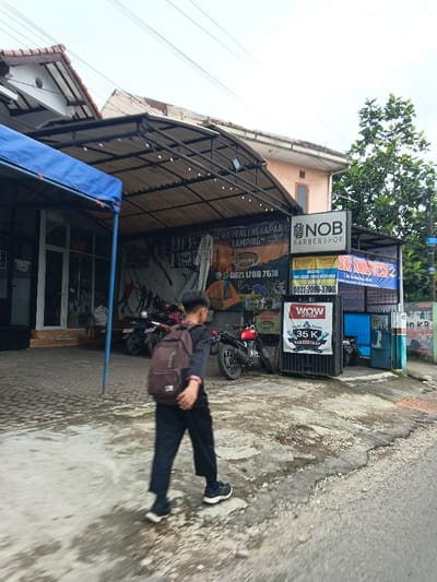 A young person with a backpack walks in front of a business area. Several motorcycles are parked near an open building with a camping supply banner that reads "SEWA PERLENGKAPAN CAMPING." The building next door has a "NOB BARBERSHOP" sign, along with smaller signs advertising pricing and services. A green metal gate is next to the barbershop. There are trees and utility poles in the background.